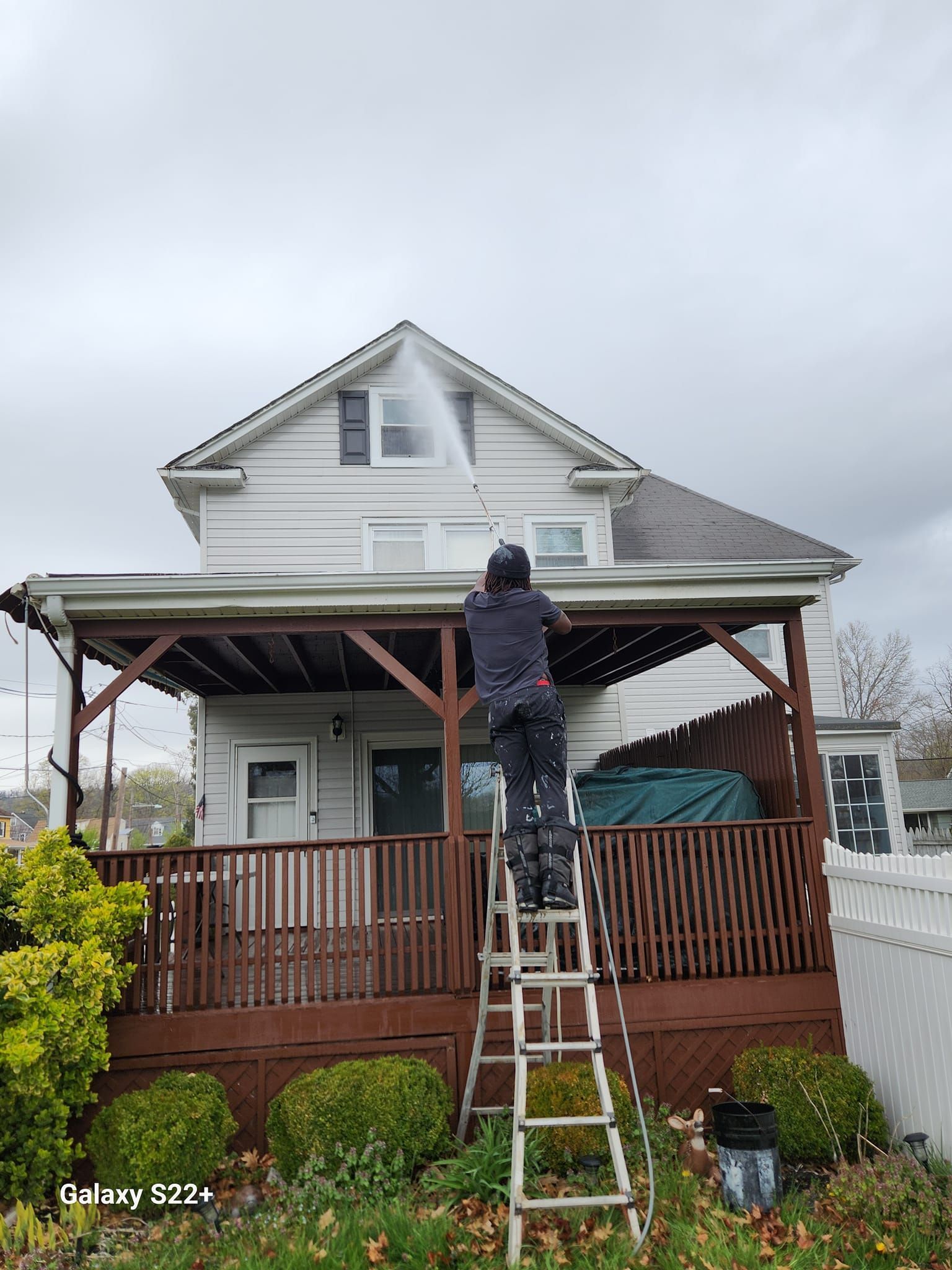 A man is standing on a ladder cleaning the roof of a house.