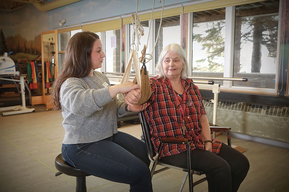 A woman is helping an elderly woman in a chair with a sling.