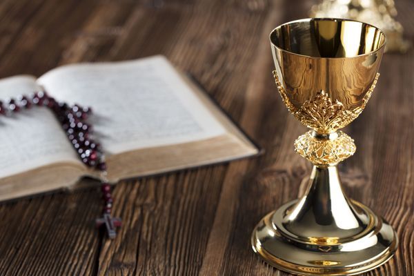 A golden chalice sits on a wooden table next to an open bible and a rosary