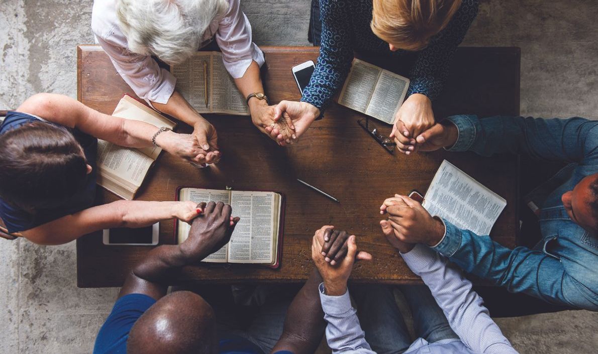 A group of people are sitting around a table with their hands folded in prayer.
