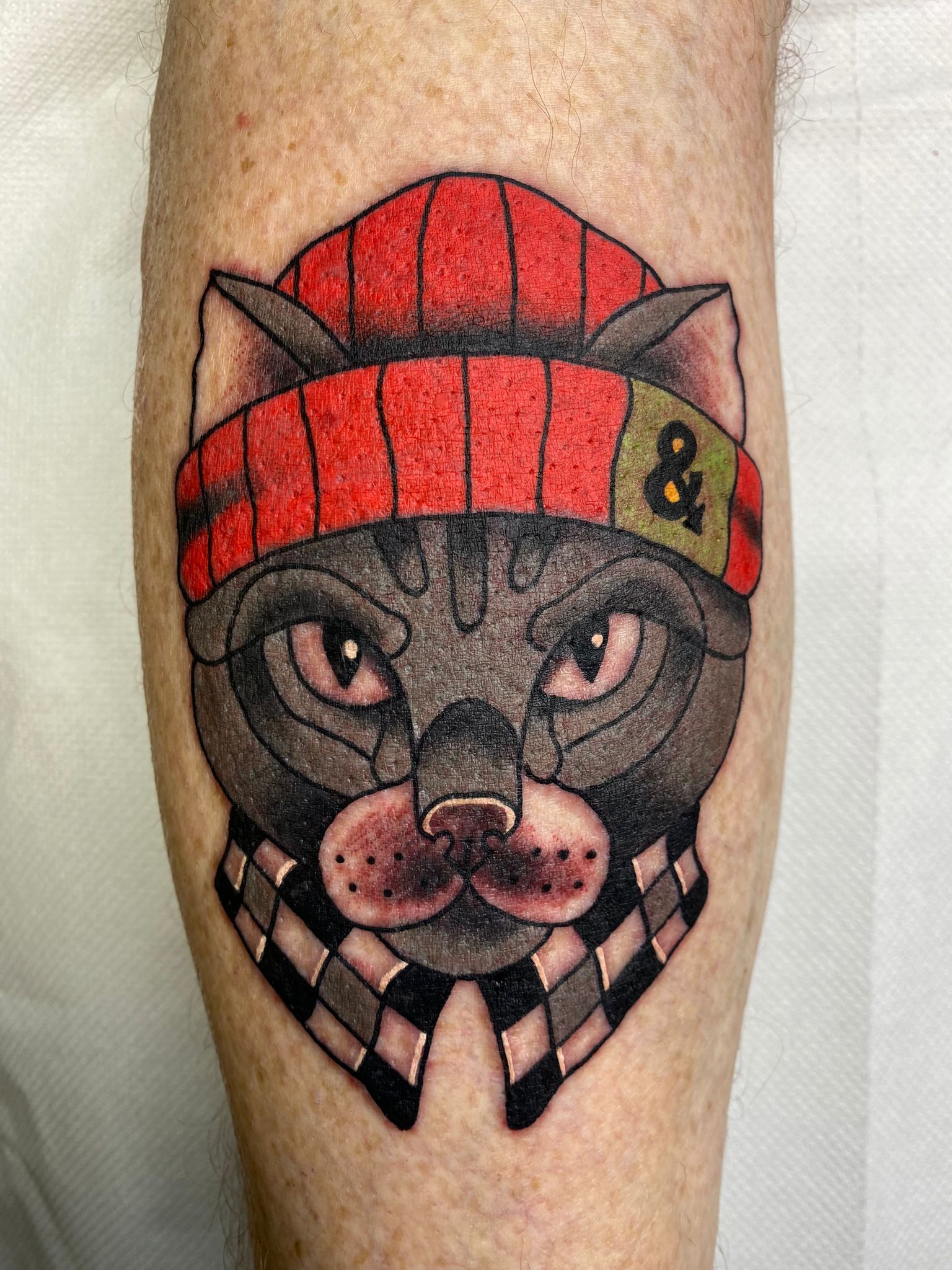 A tattoo of a cat wearing a hat and scarf on a leg.