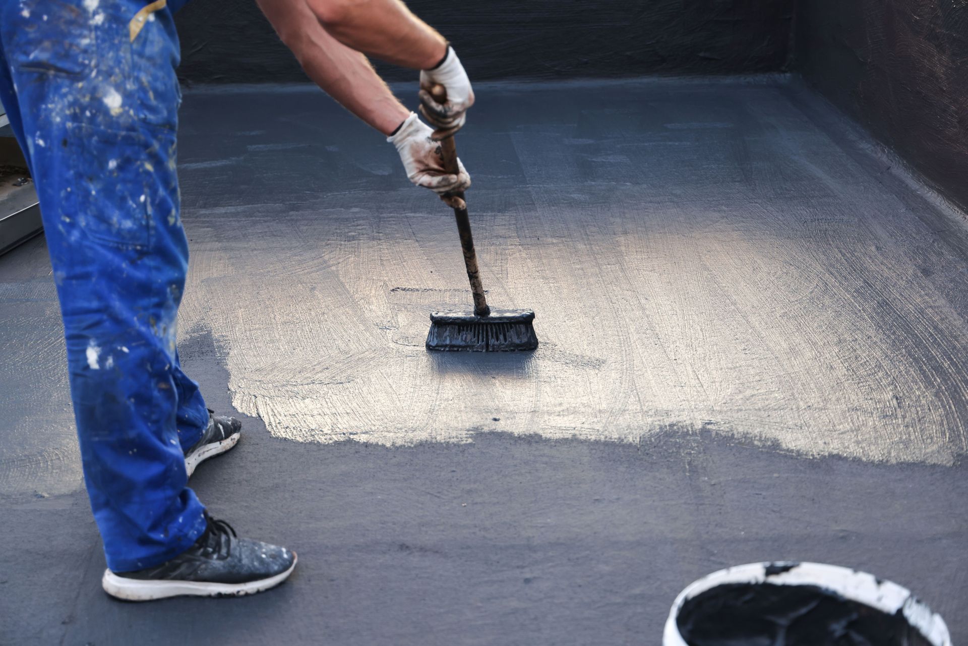 Person applying black sealant to a flat roof with a brush, wearing blue overalls.