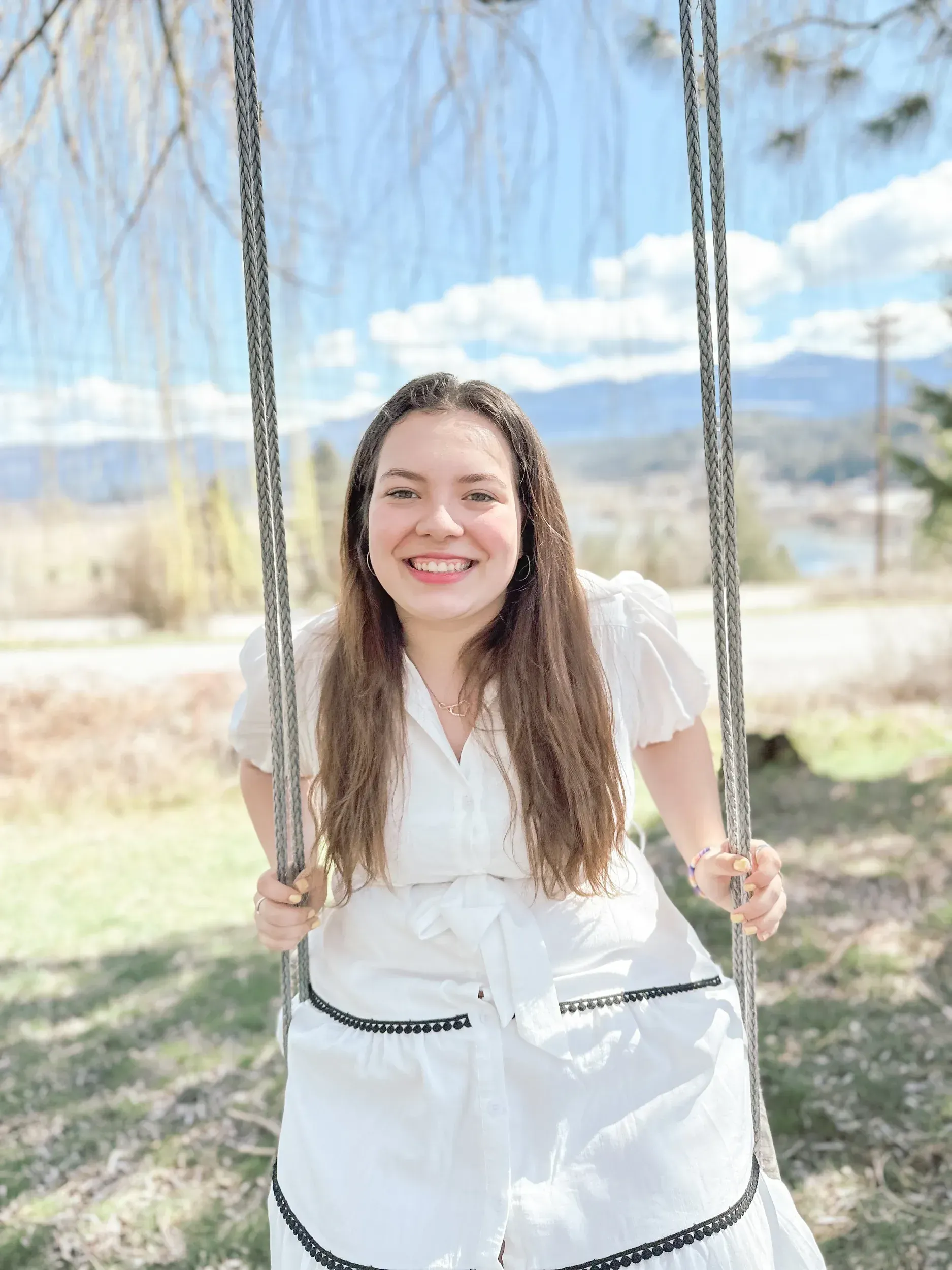 Woman smiling on a swing outdoors, wearing a white dress with mountains in the background.