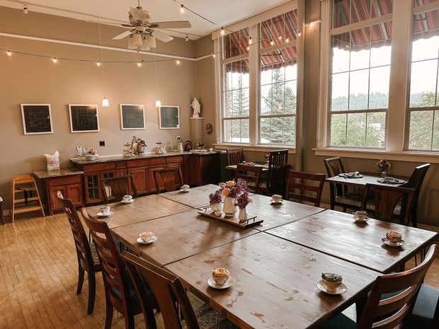 A dining room with wooden tables, chairs, and large windows, featuring a light blue wall, ceiling fan, and wooden floor.