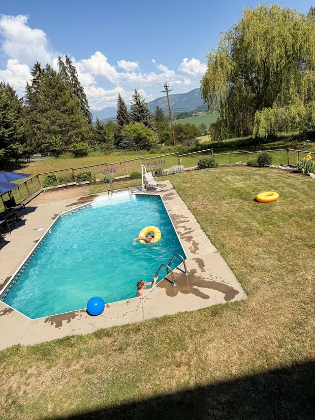 A rectangular swimming pool surrounded by a concrete deck and a lush green lawn, set against a backdrop of trees and hills.
