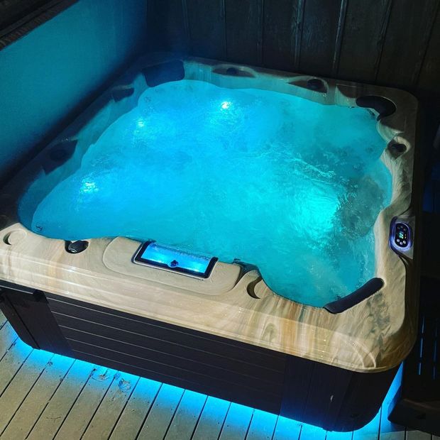 A square, marbled-finish hot tub with bright blue interior and base lighting, located on a wooden deck.