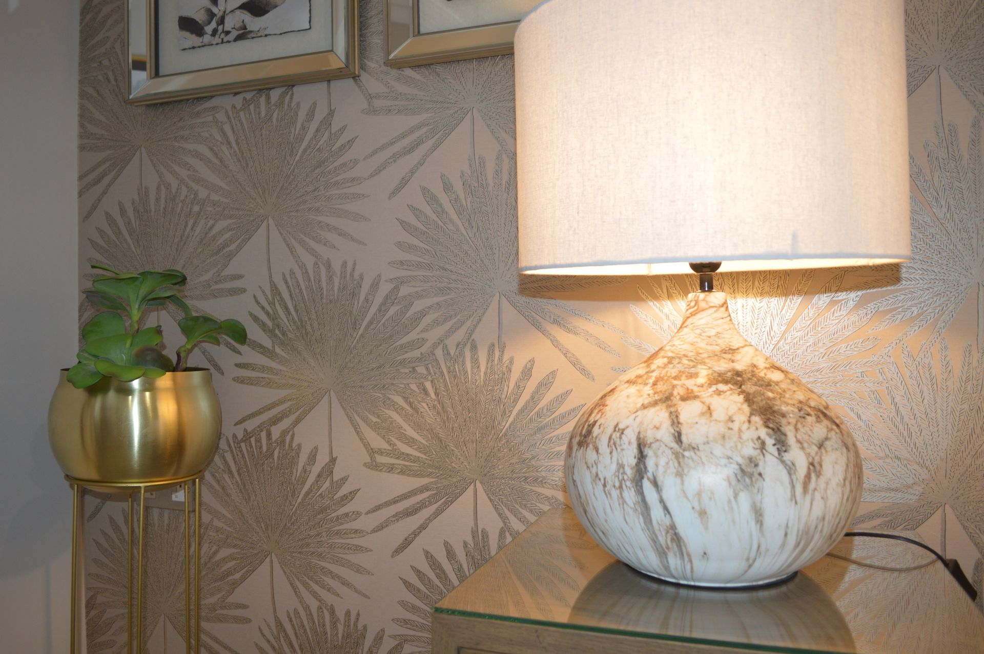 A lamp is sitting on a table in front of a wall with palm leaves on it.