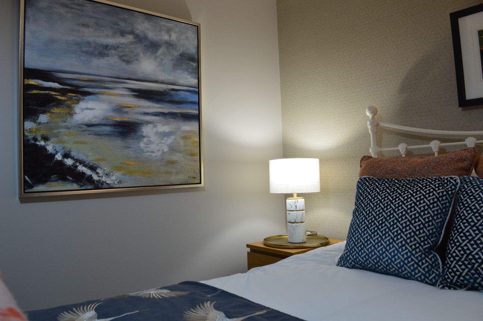 A bedroom with a bed , nightstand , lamp and painting on the wall.