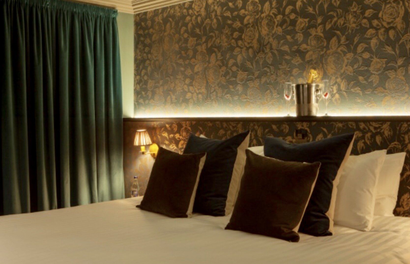 A hotel room with a bed , pillows , a lamp and a bottle of champagne.