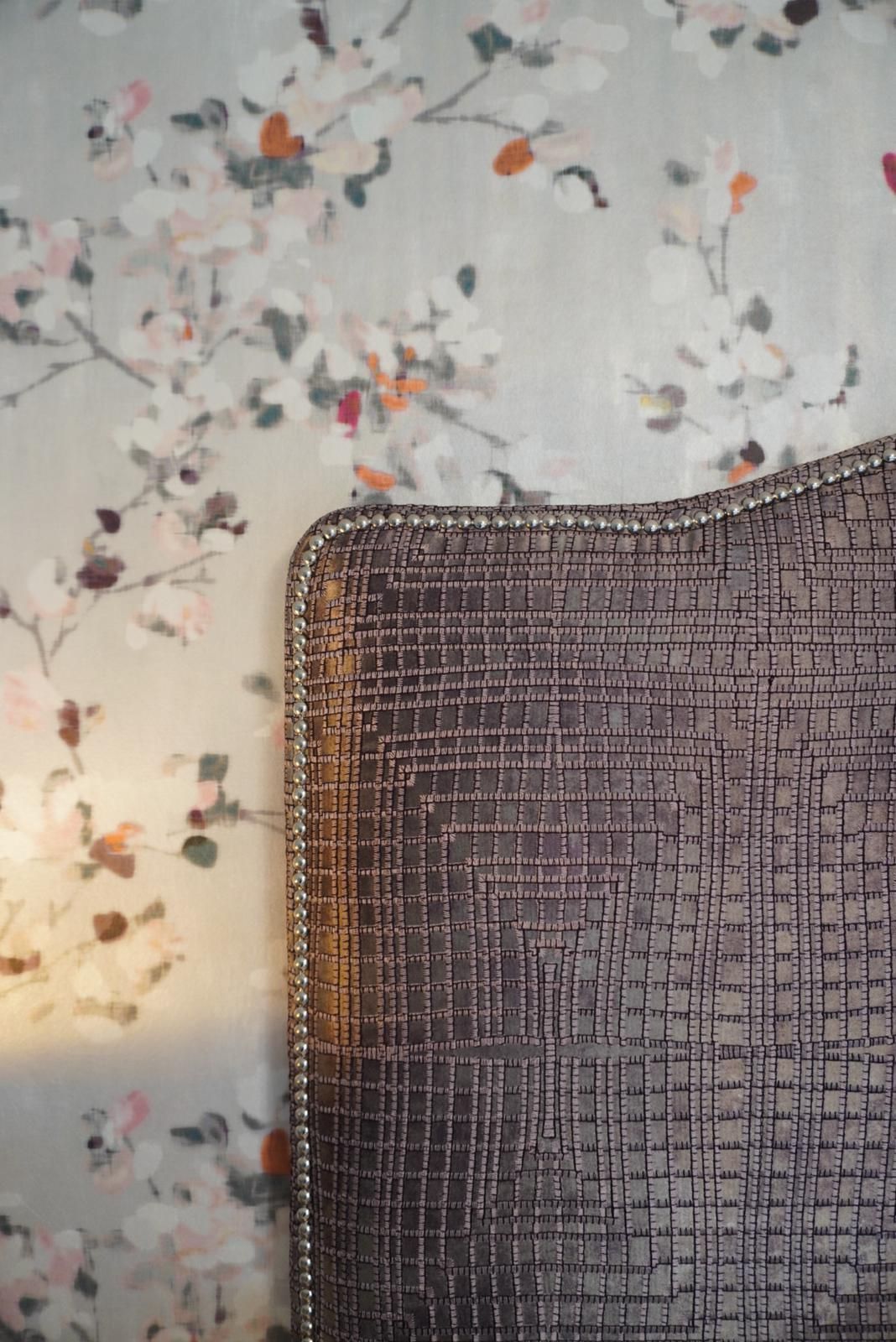 A close up of a pillow on a bed in front of a floral wallpaper.
