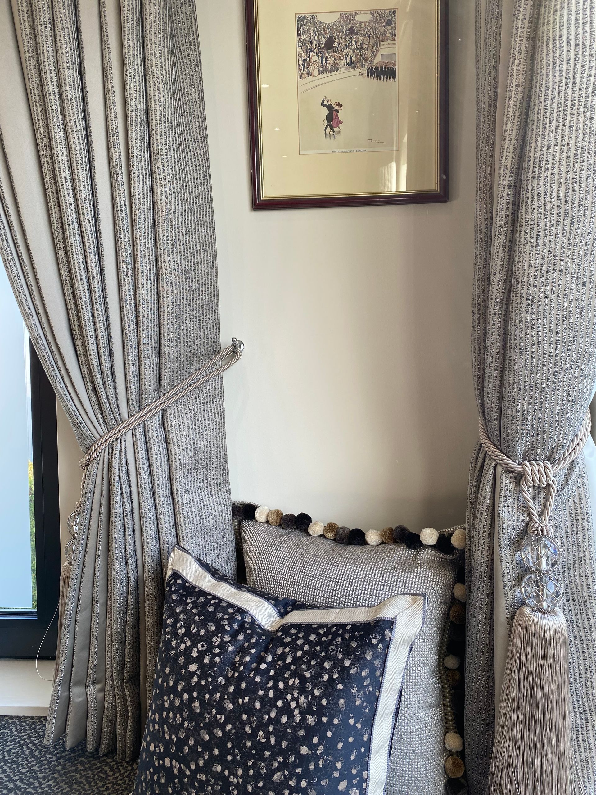A corner of a room with curtains and pillows and a picture on the wall.