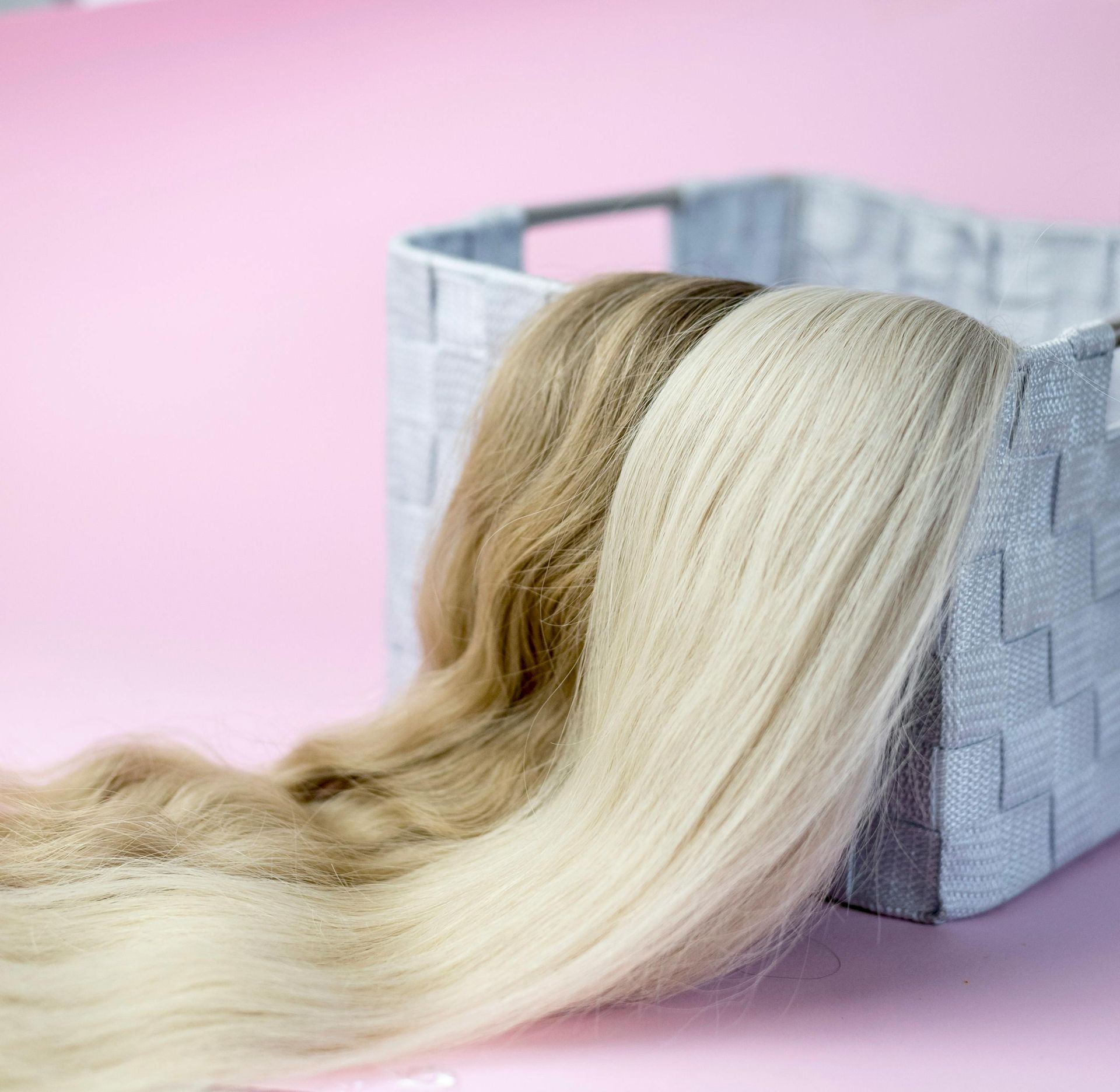Blonde and light brown hair extensions spilling from a gray basket on a pink background.