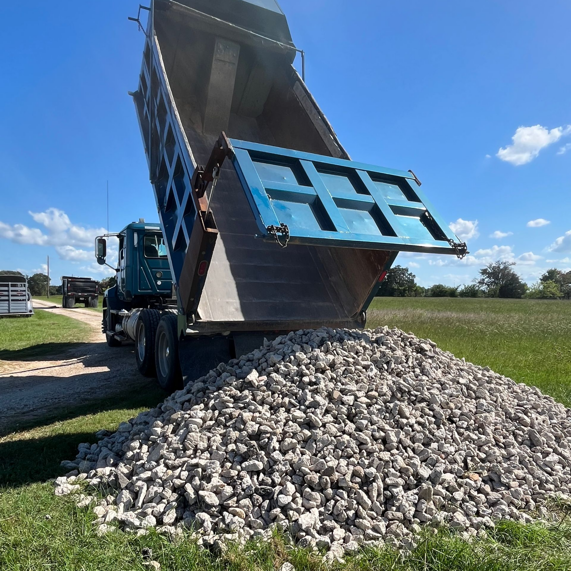 Dump truck unloading gravel onto a pile in a grassy field on a sunny day.