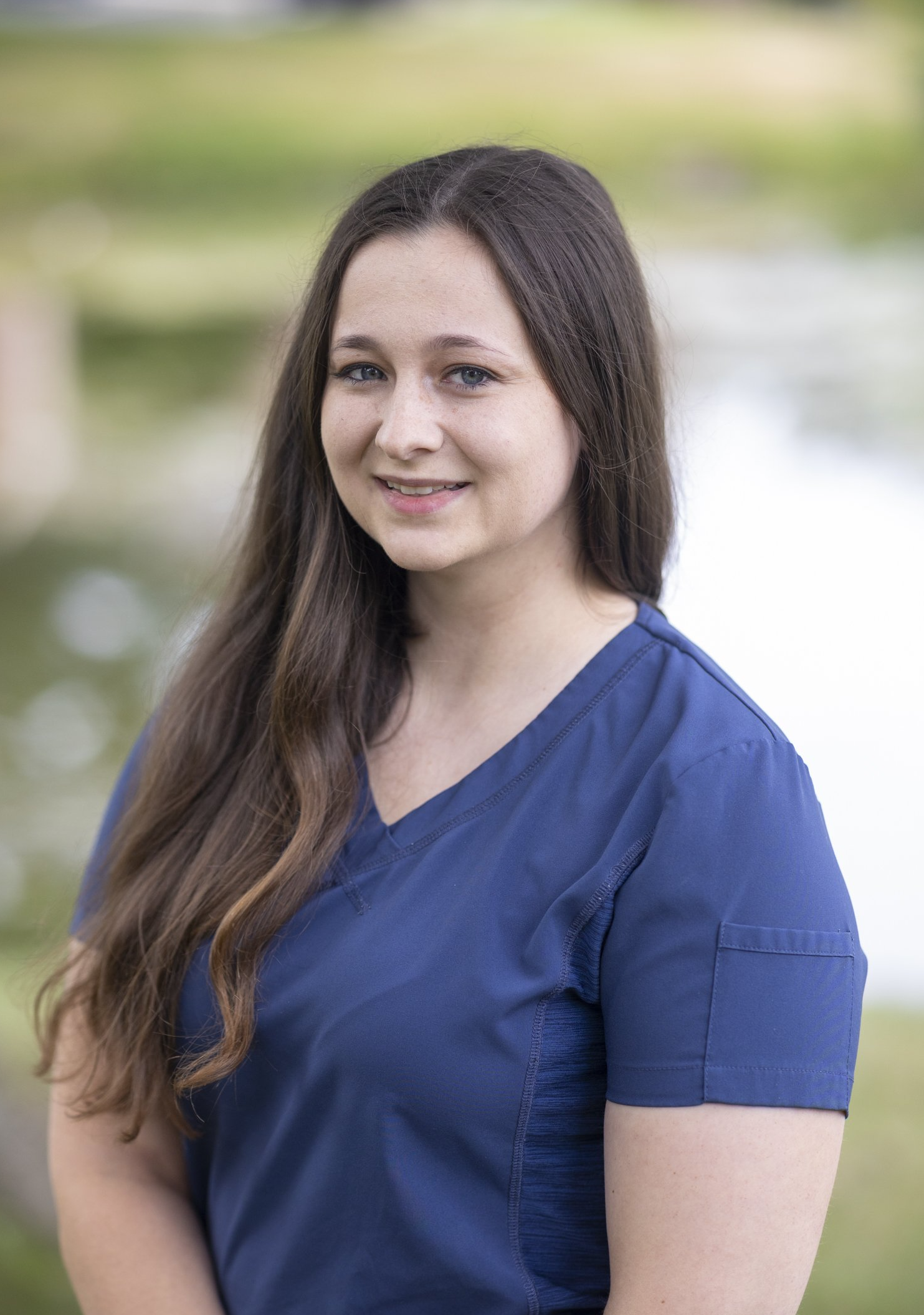 A woman in a blue scrub top is smiling for the camera at Advanced Dermatologic Surgery in Overland Park, KS