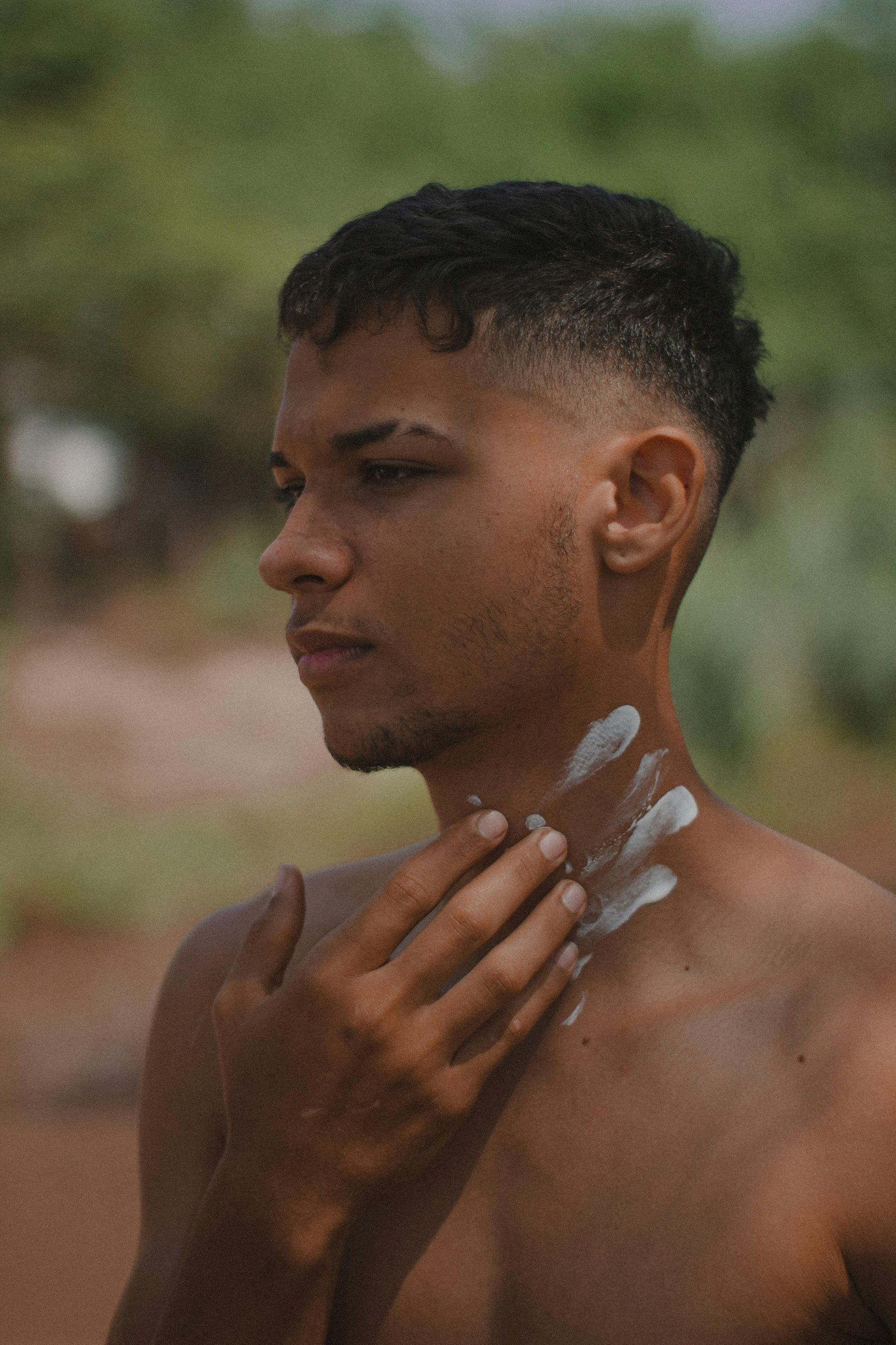 A person applying sunscreen to their neck in an outdoor, blurred natural setting.