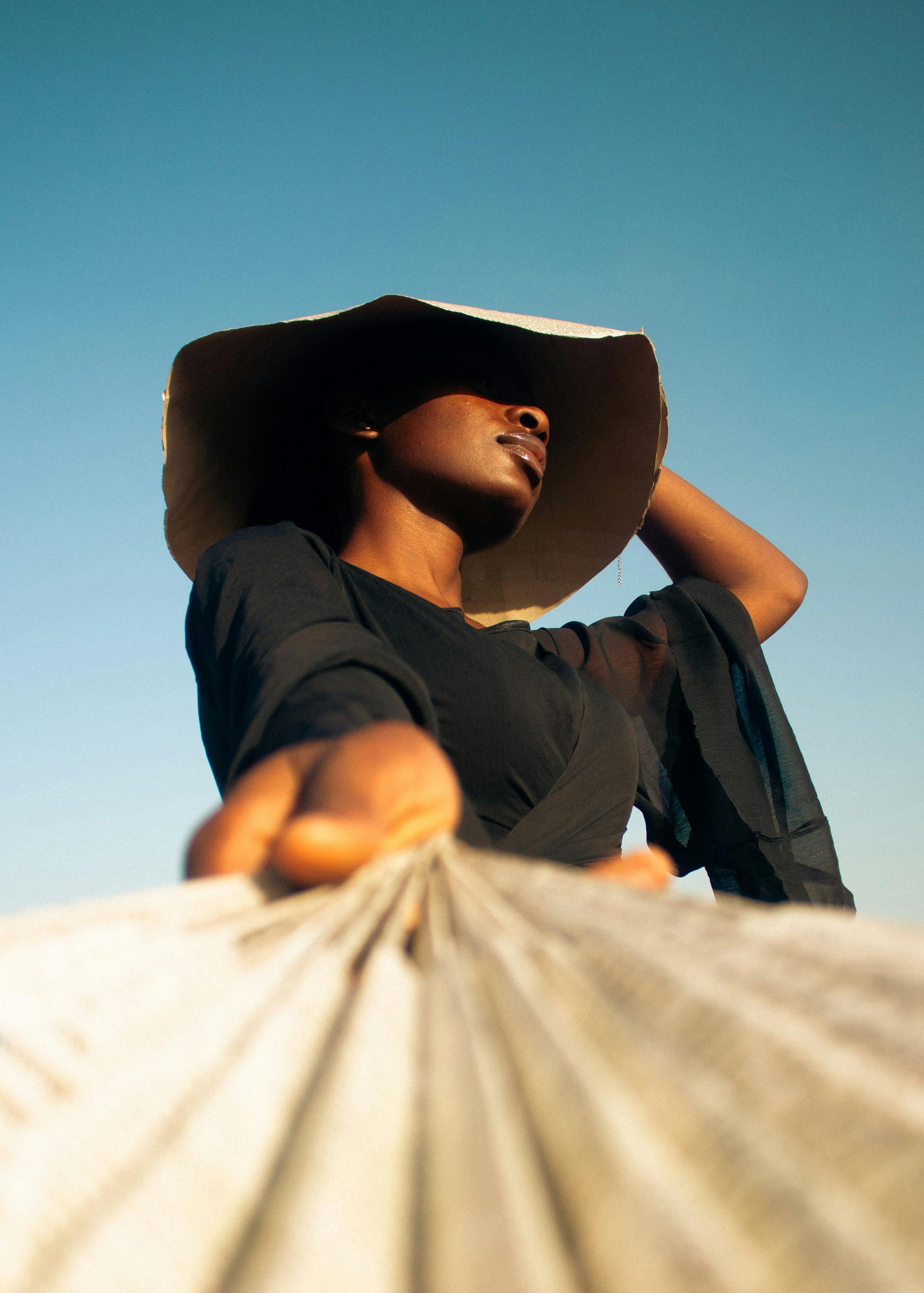 A person in a dark top and wide-brimmed hat holds a textured fabric against a clear blue sky.
