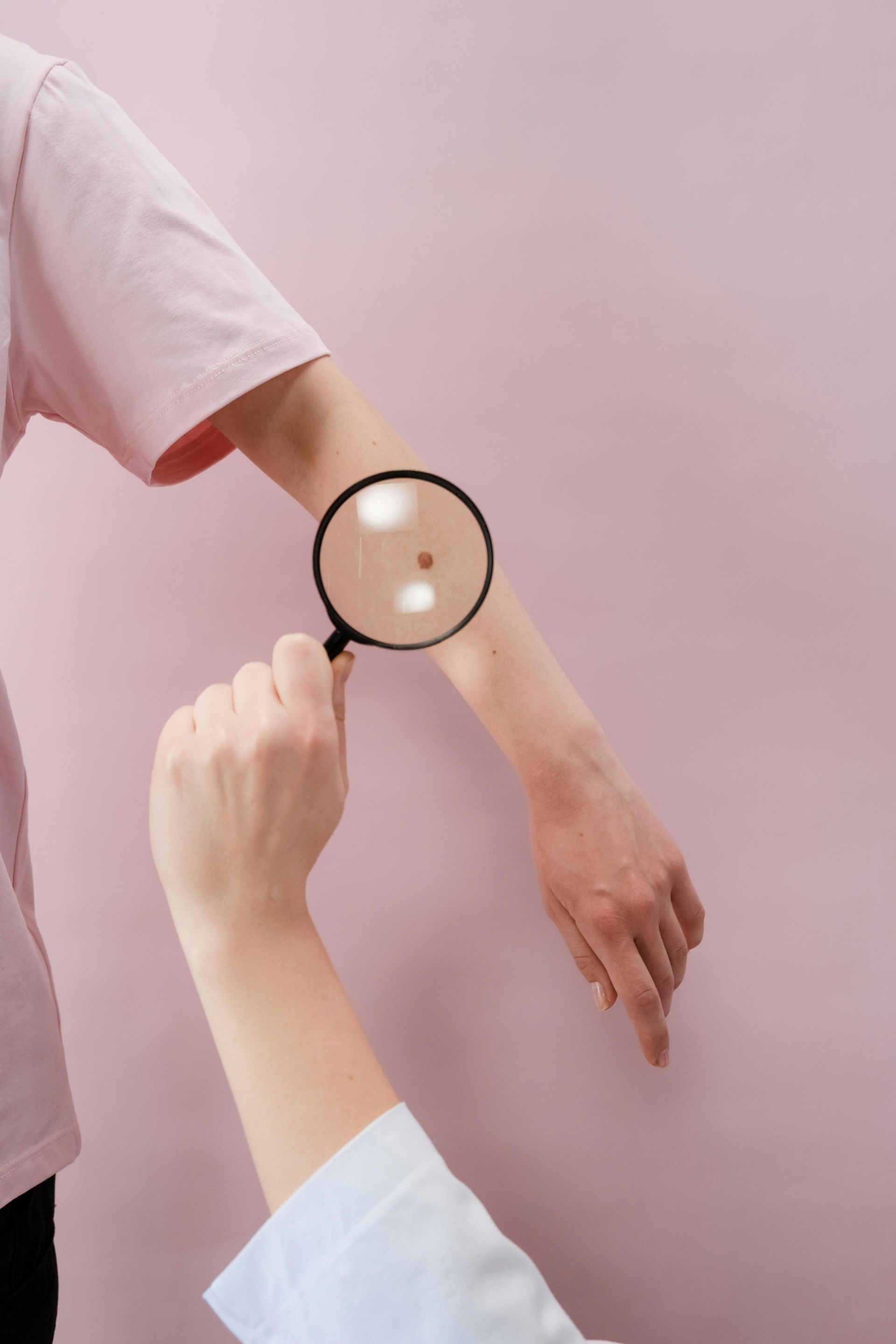 Person examining a mole on an arm with a magnifying glass, pink background.