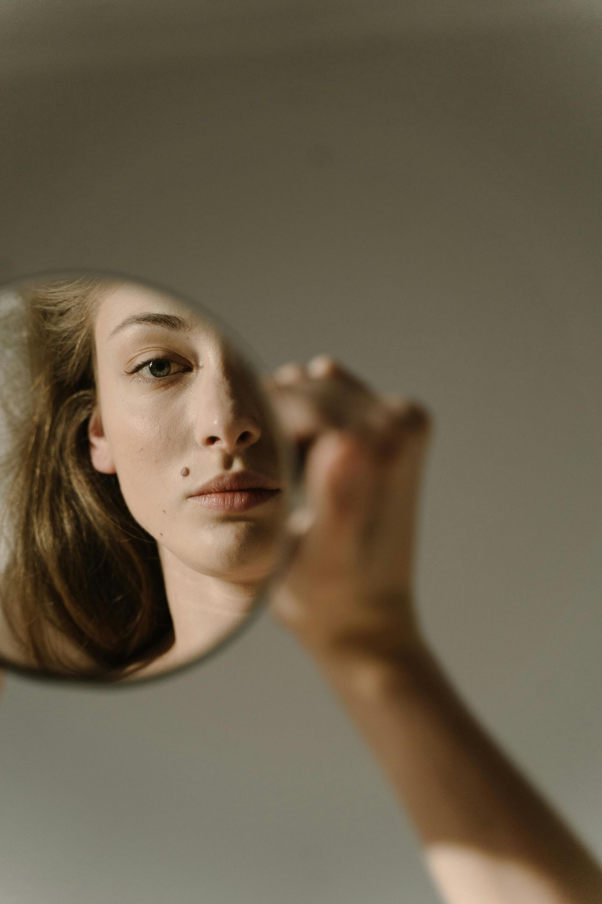 Woman looking in a small round mirror; hand in focus, face and upper body visible. Light on beige background.