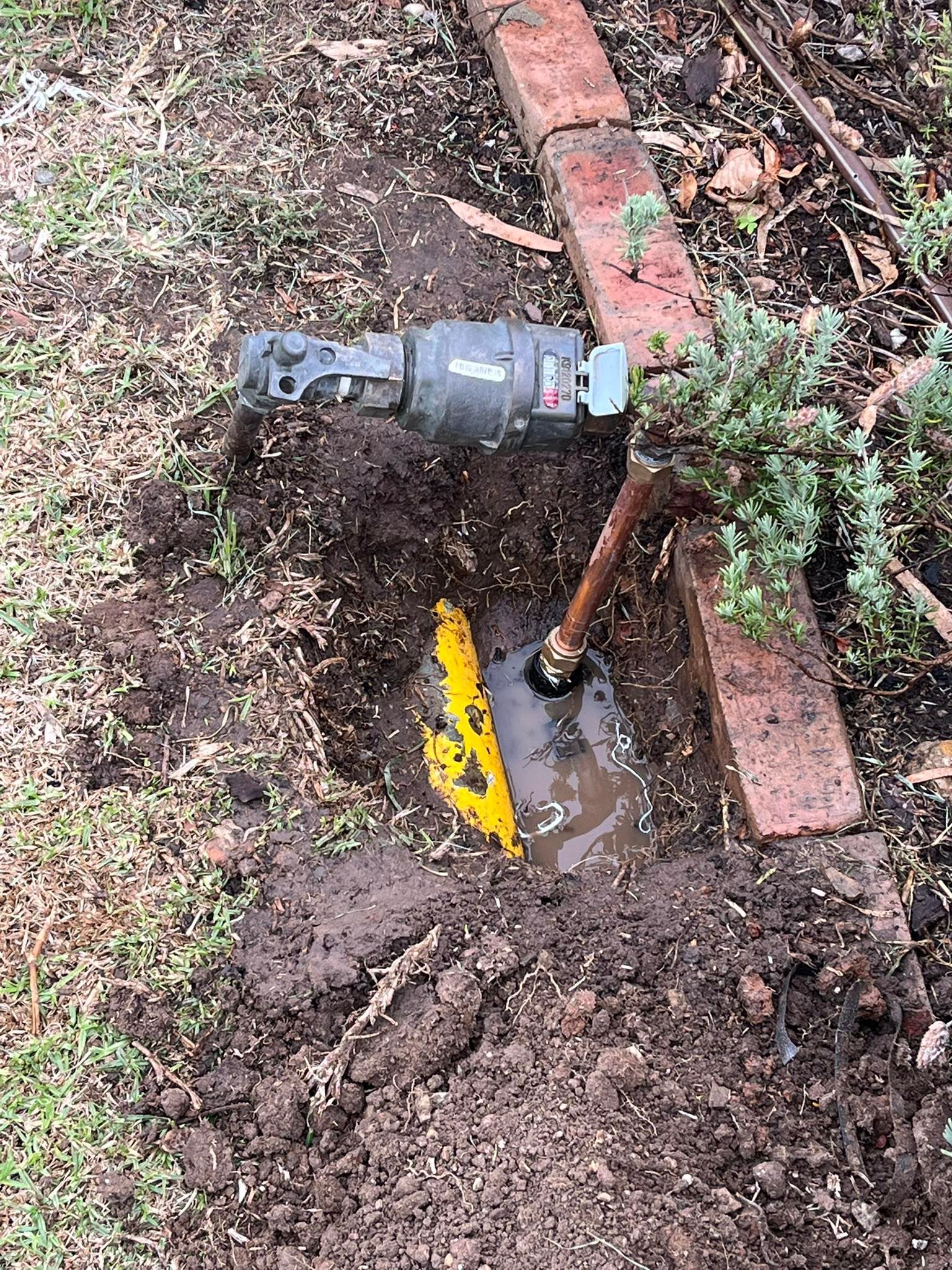 A residential water meter and copper pipe located in a muddy hole next to a brick border in a garden bed — Leak & Co in Zuccoli, NT 