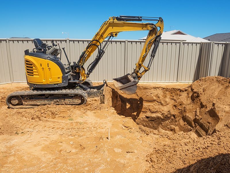 Yellow excavator digging a hole in sandy soil; a white fence and blue sky are in the background.