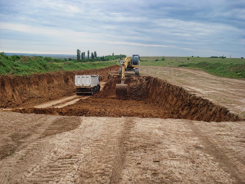 An excavator loads dirt into a dump truck in a construction zone, with a cloudy sky overhead.