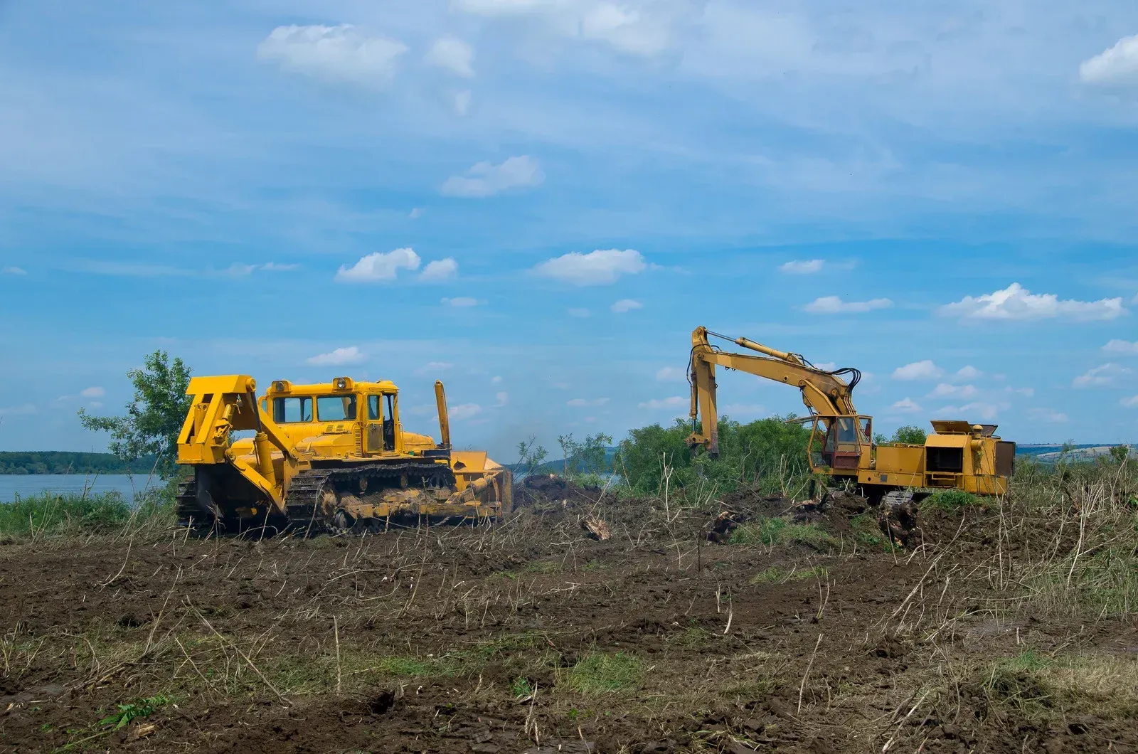 Yellow bulldozer and excavator clearing a brown field under a blue sky with scattered clouds.
