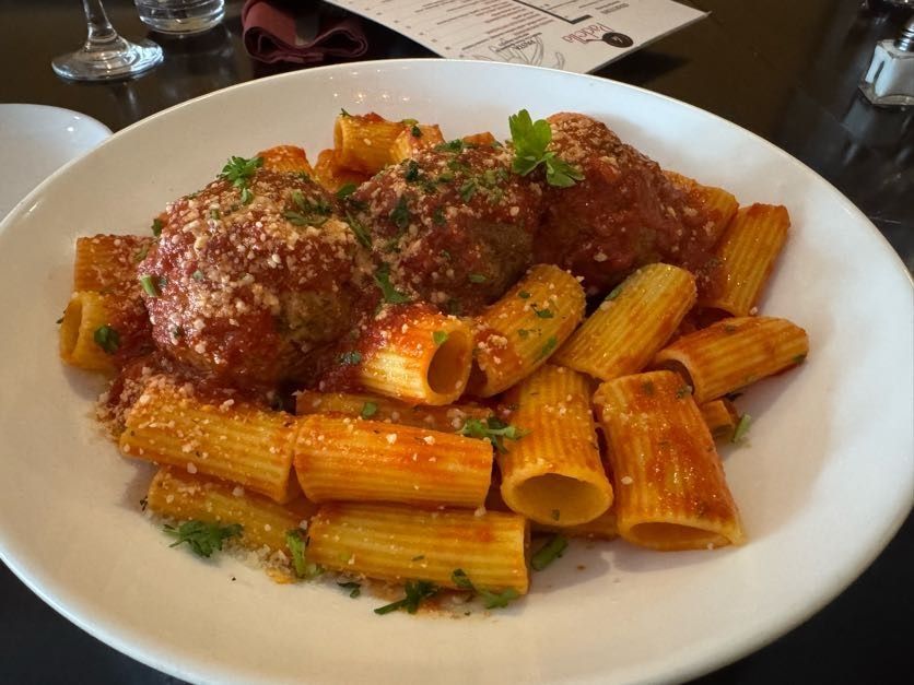 Rigatoni pasta with meatballs and tomato sauce on a white plate, garnished with parsley.