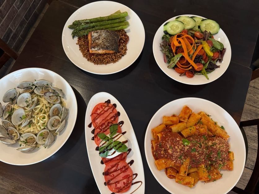 Five plated dishes on a dark table: clams, salmon with asparagus, salad, sliced meat, and pasta.