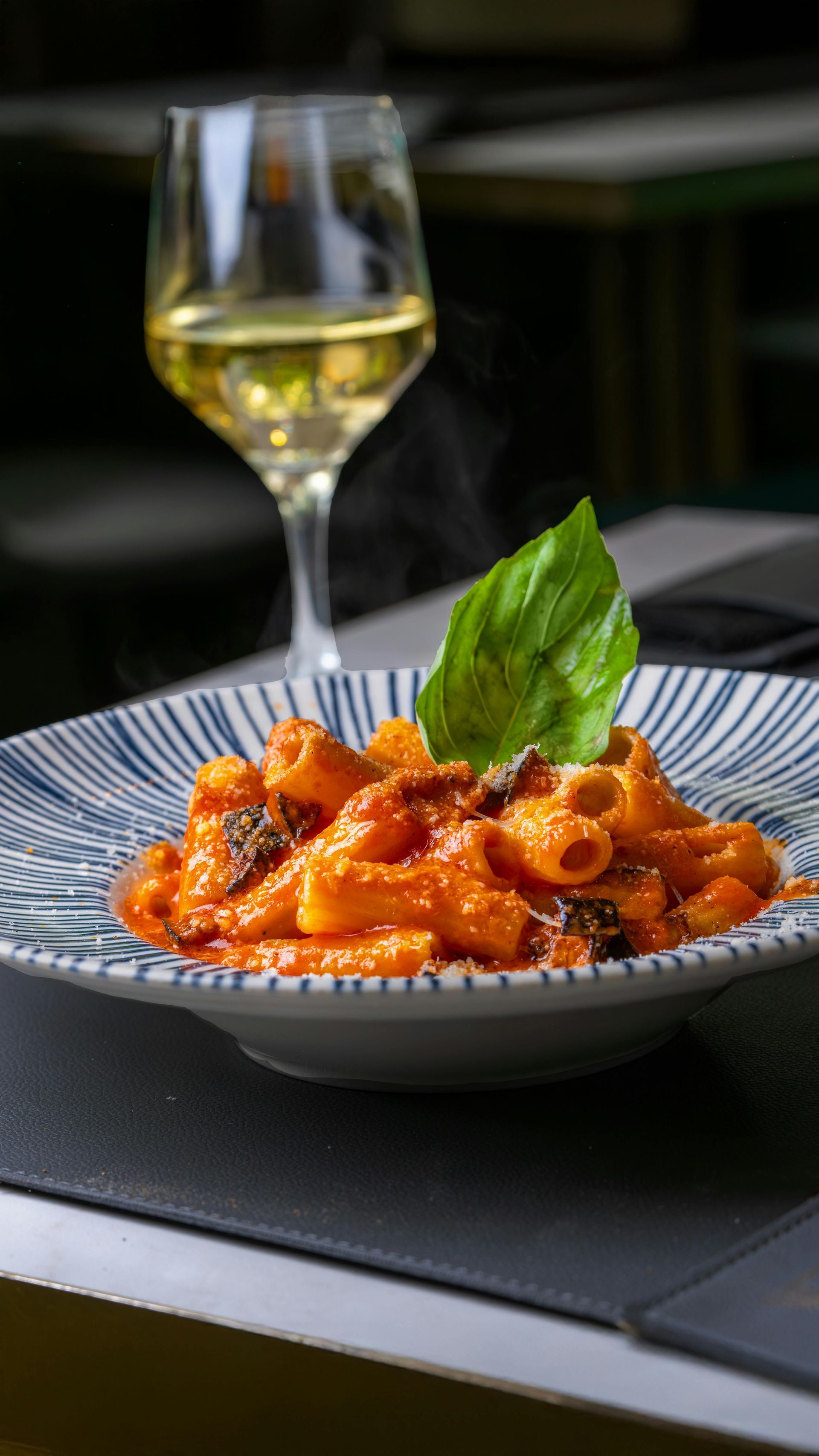 Pasta in red sauce with basil on a striped plate, beside a glass of white wine on a table