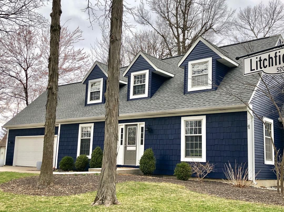 A blue house with a gray roof and white trim is surrounded by trees.