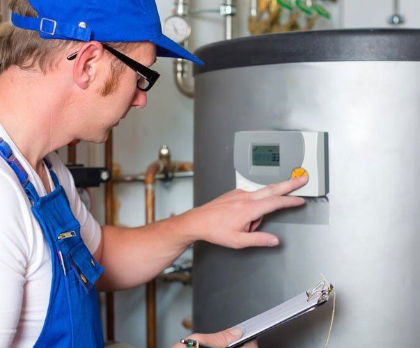 A man wearing a blue hat and overalls is working on a water heater while holding a clipboard.