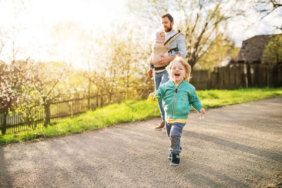 A man is carrying a baby in a carrier and a little girl is running down a road.