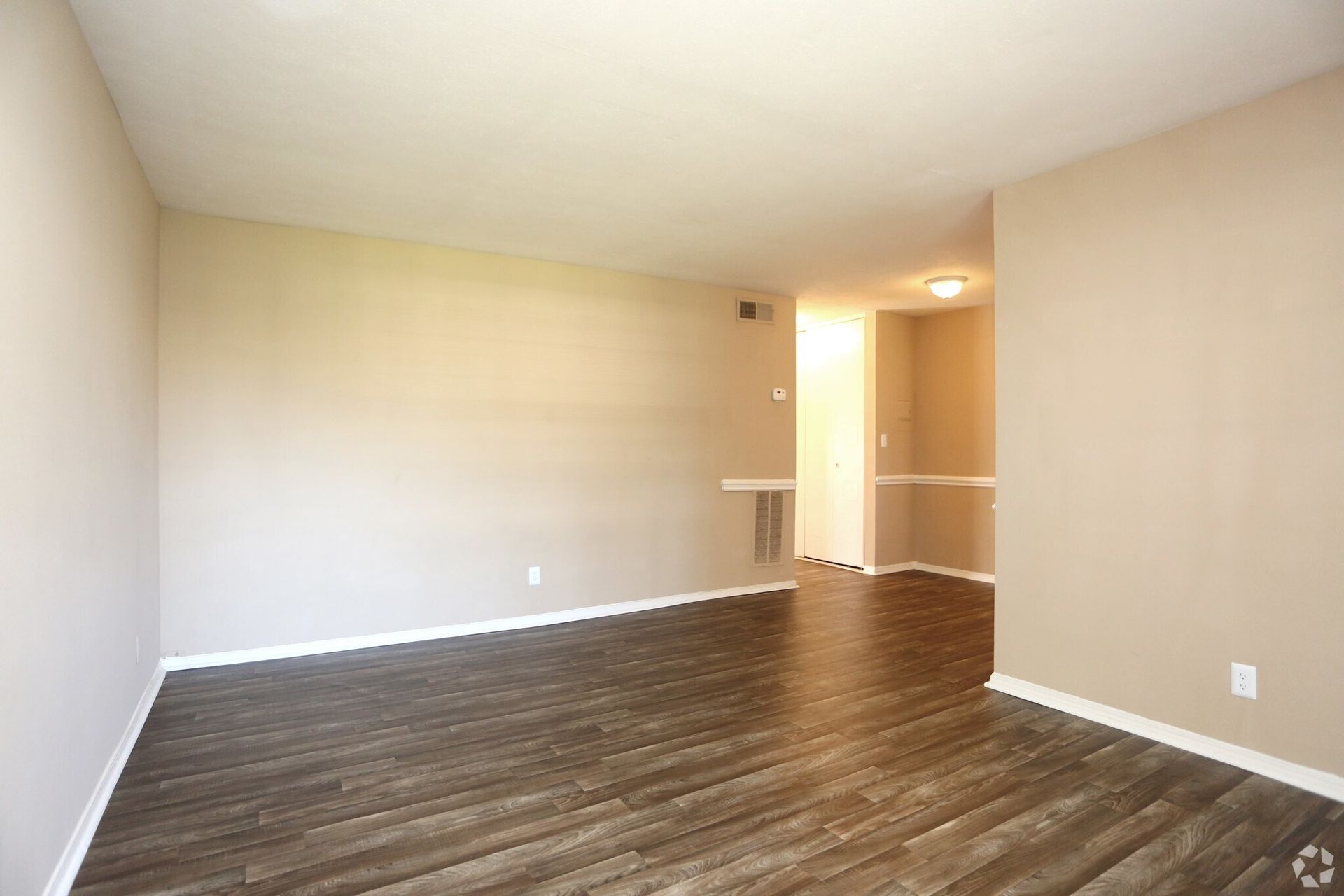 Empty living room with wood-look flooring and beige walls. Doorway to a lit hallway.