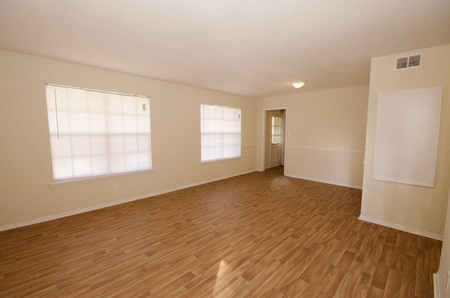 Empty room with wood-look flooring, beige walls, two windows with blinds, and a doorway.