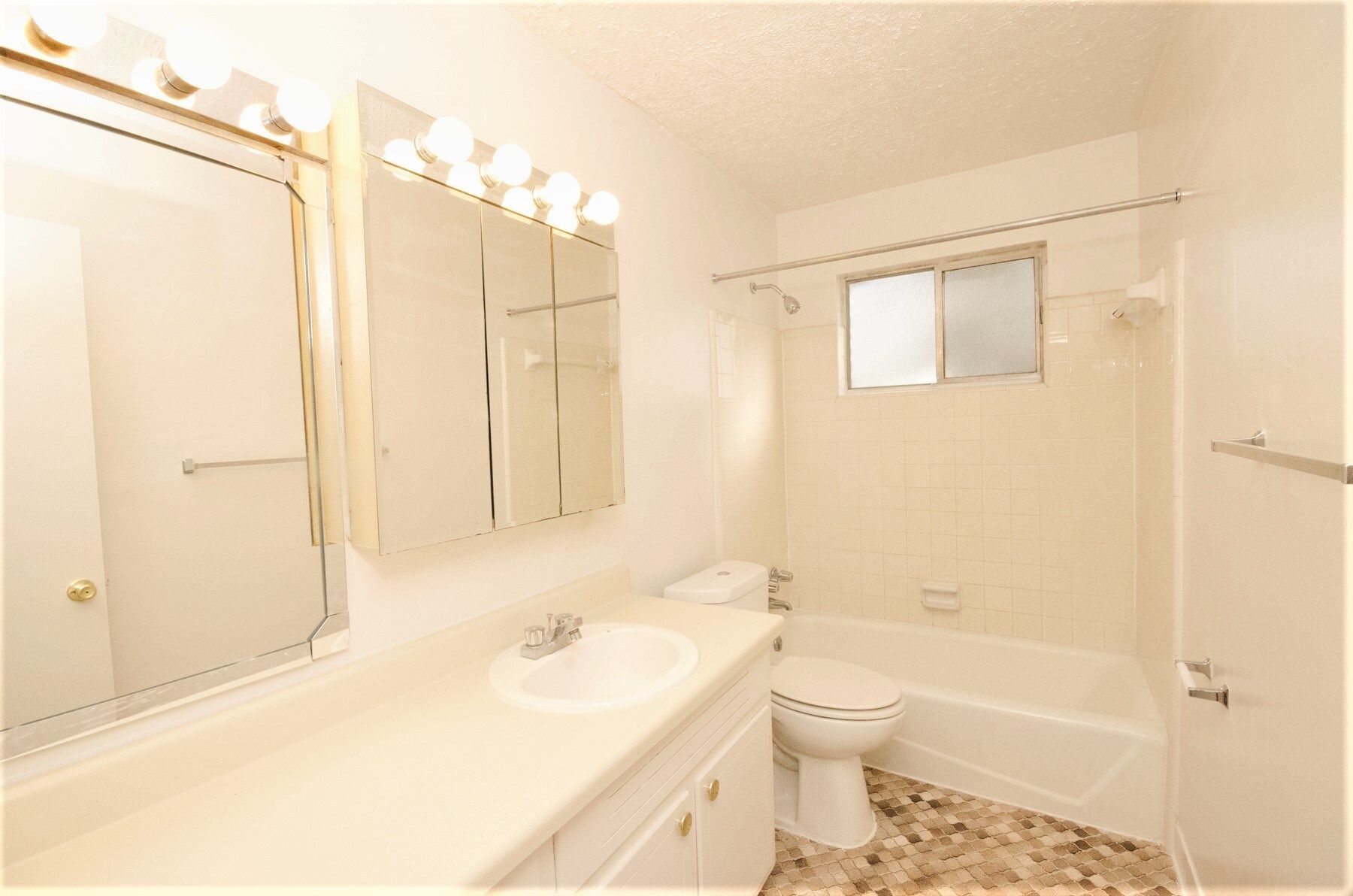 Bathroom with a white vanity, toilet, and bathtub. Beige tiled floor, window, and bright lights.