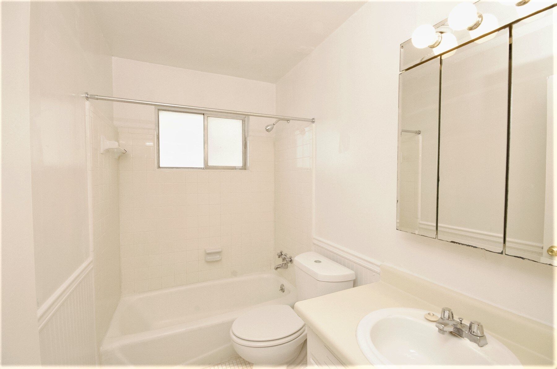 White bathroom with a bathtub, toilet, sink, and mirror with three overhead lights.