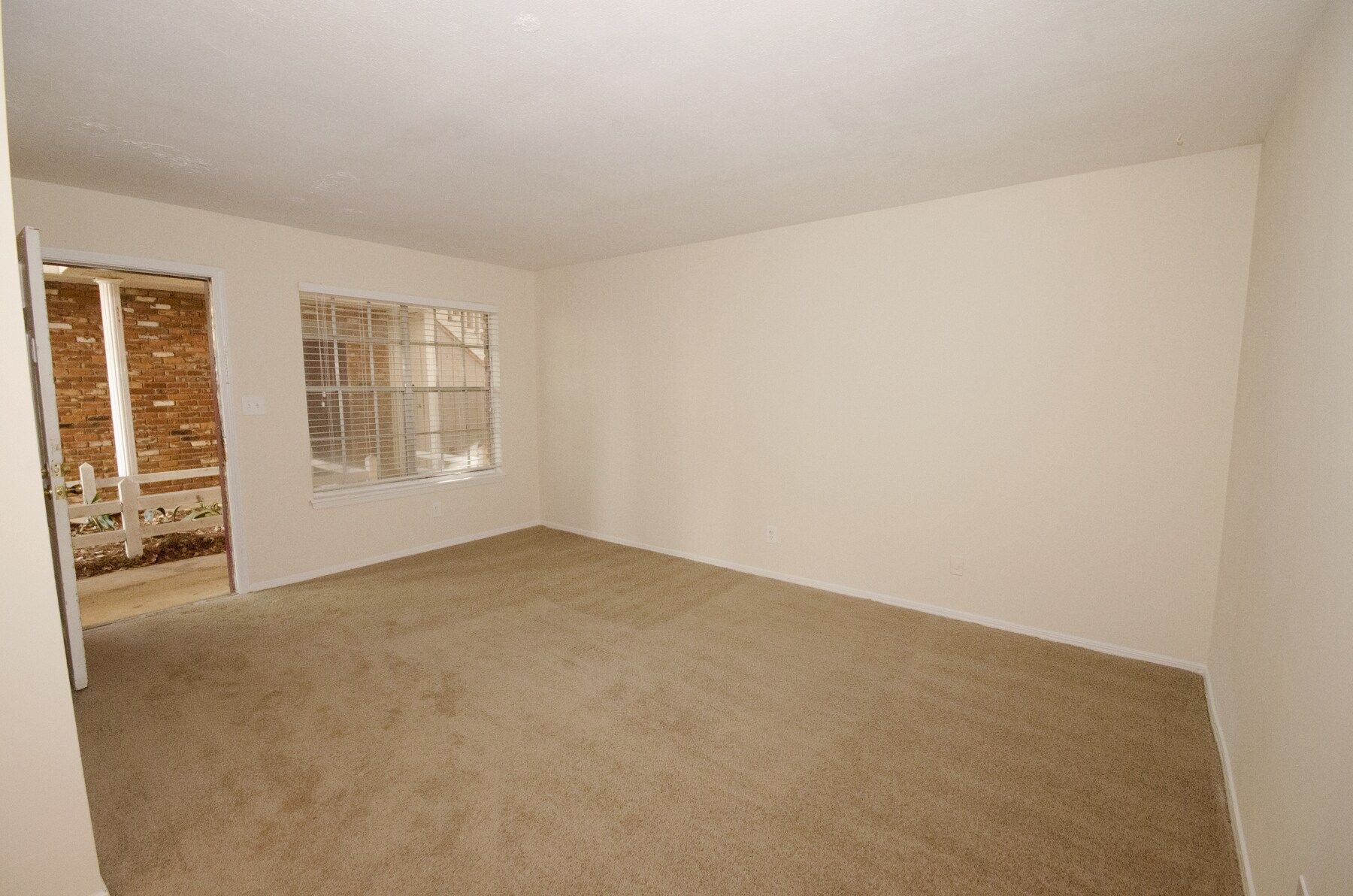 Empty room with beige carpet and walls; doorway on the left, window with obscured view on the back wall.
