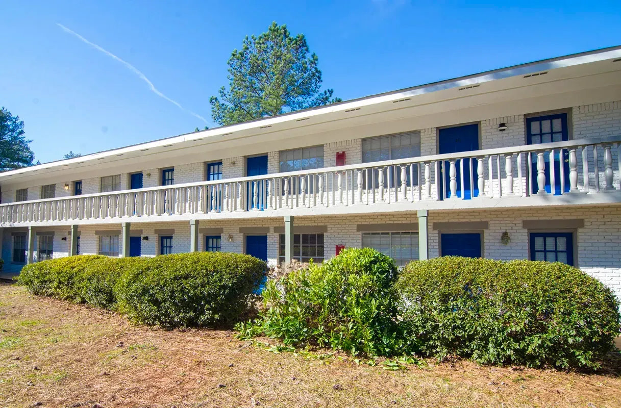 Two-story white brick apartment building with blue doors and a wooden balcony, bushes in front.