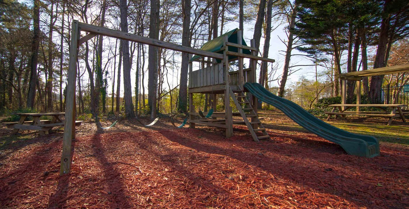 Playground in a wooded area with swings, slide, and wooden structure; ground covered in wood chips.