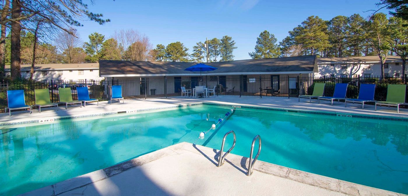 A swimming pool with blue and green chairs. A building is behind the pool with trees in the background.