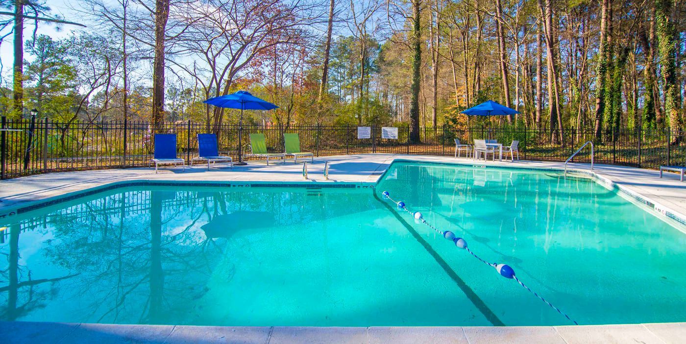 A clear, turquoise swimming pool with blue umbrellas, chairs, and a treeline in the background on a sunny day.