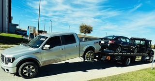 A silver truck is towing two cars on a flatbed tow truck.