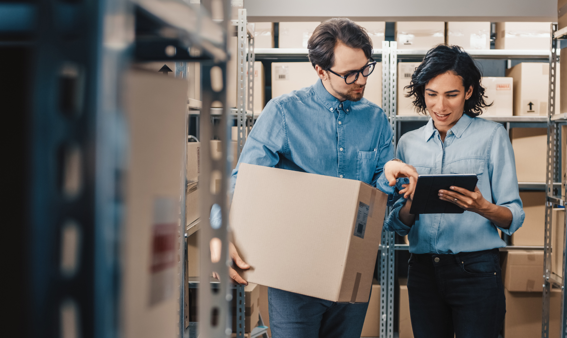 Two people in warehouse, man holding box, woman looking at tablet, discussing.