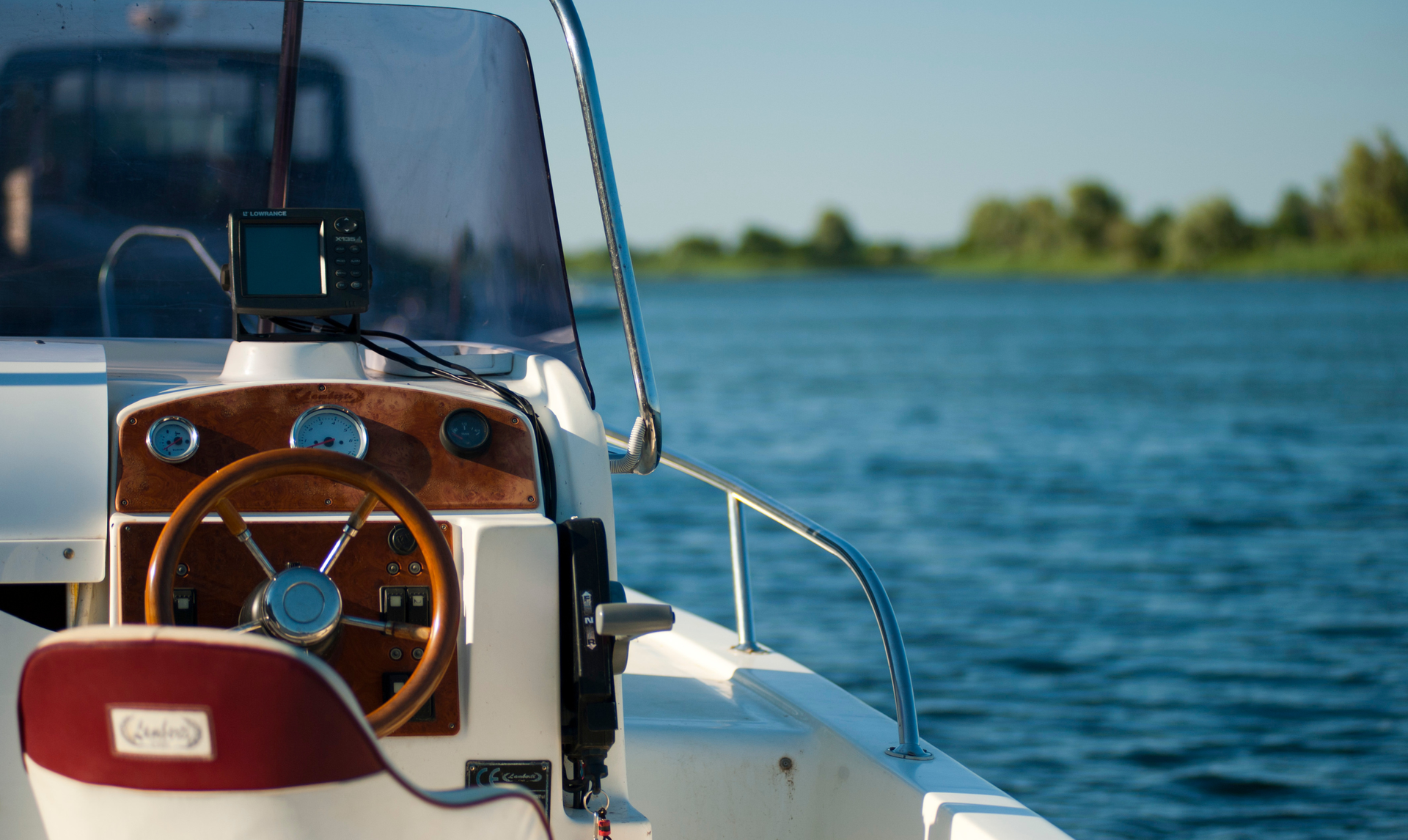 Boat cockpit with wooden steering wheel, open water, and treeline in background.