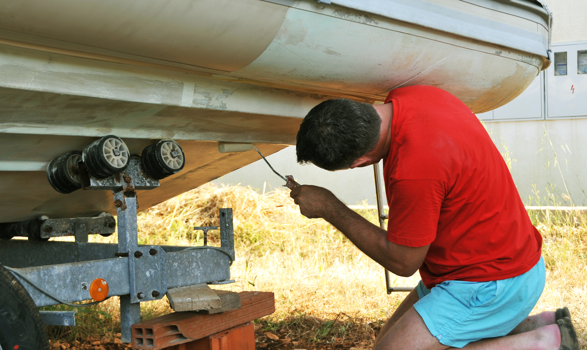 Man kneeling, inspecting the underside of a boat on a trailer, holding a tool; outdoors.