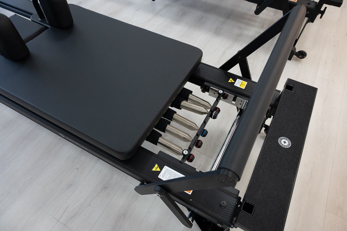 Black Pilates reformer machine, springs visible, resting on a light wood floor.