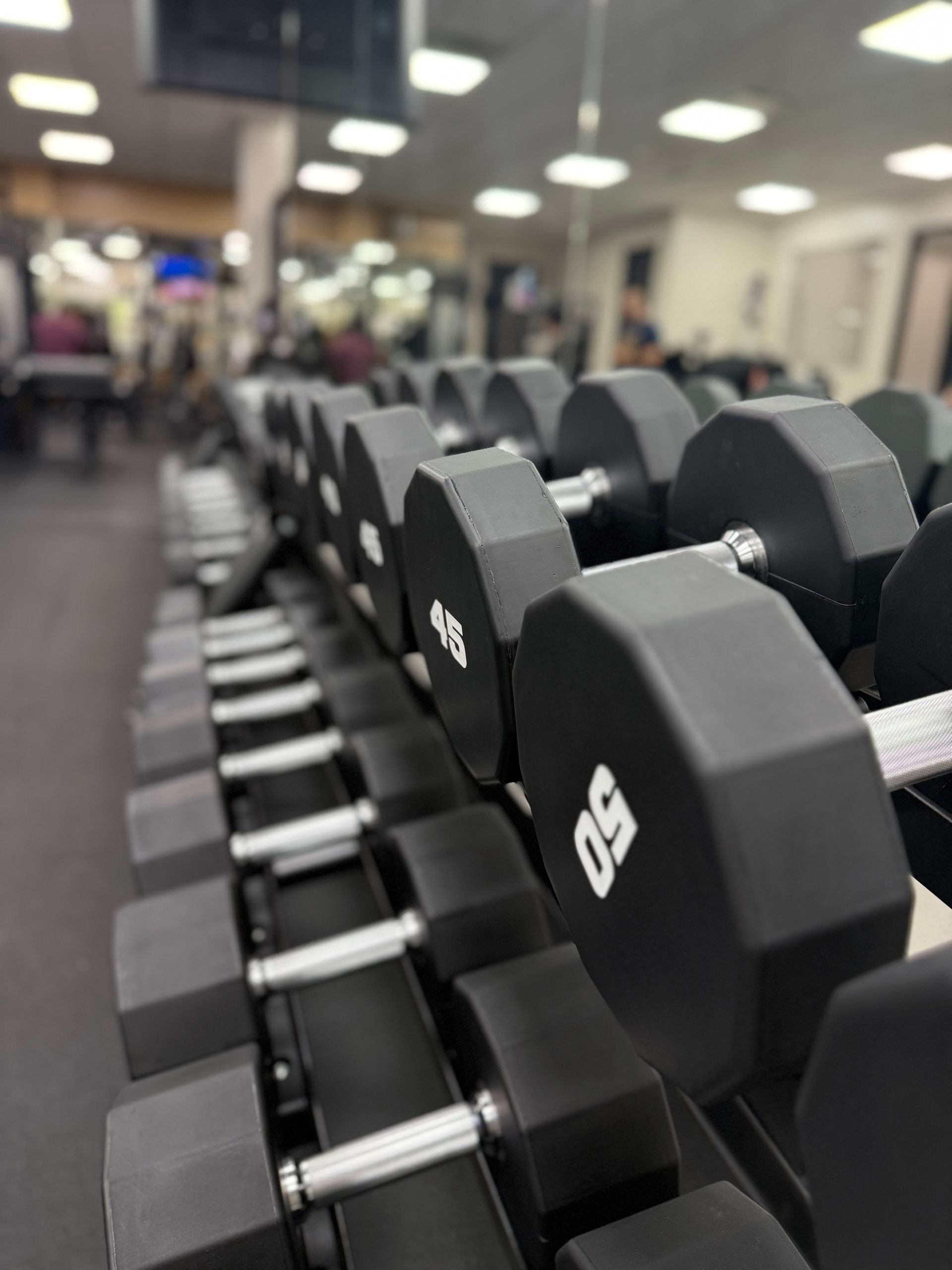 Rows of dumbbells in a gym, the nearest pair are 50lbs, black and chrome, with other equipment blurred in the background.