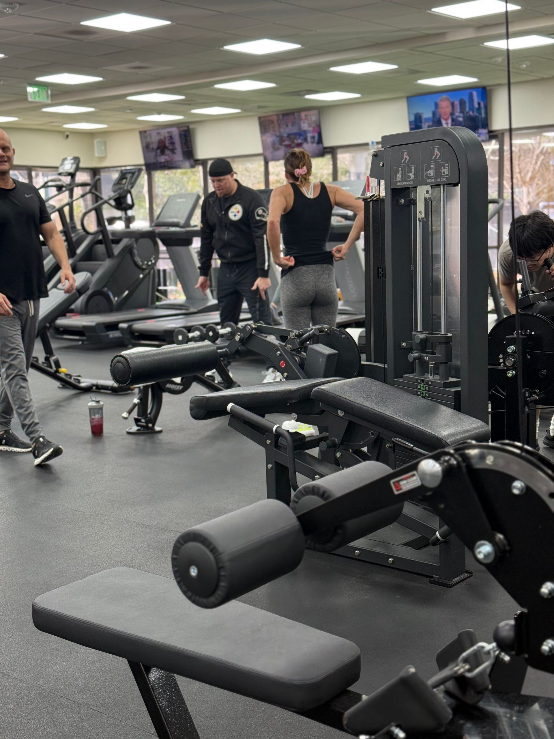 Gym interior. People working out on various machines: treadmills, weight machines. Black and gray equipment, fluorescent lighting.