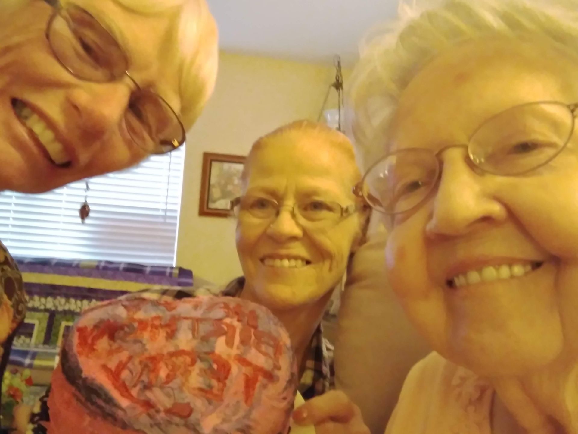 Three older women are posing for a picture, and one of them is holding a heart that says happy birthday