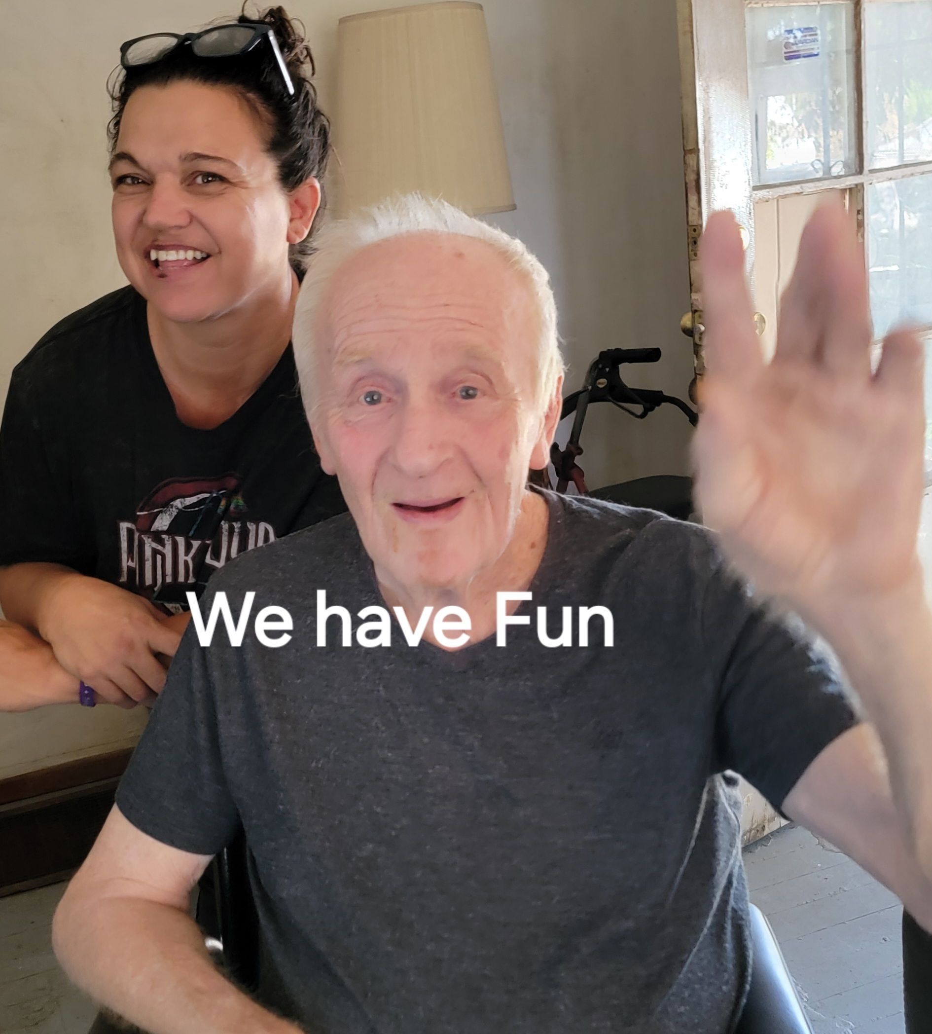 An elderly man in a wheelchair with a woman behind him that says we have fun